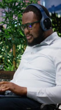 Dikey video özgüvenli yetişkin adam uzaktan çalışma için odak müzik dinliyor, bir bankta oturuyor ve çevrimiçi işler için dizüstü bilgisayar kullanıyor. Metropolitan bölgesinde moda etkinliği, modern şehir merkezi caddesi