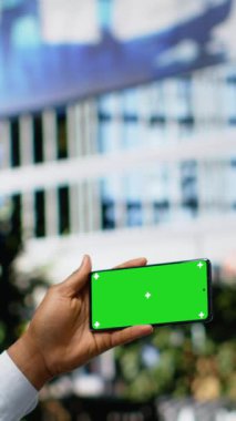 Şehir merkezindeki bankta oturan siyah bir adamın bulunduğu dikey video yeşil ekran akıllı telefon güneş ışığı altında çevrimiçi içeriği okuyup tarıyor. Modern mimari ve şehir merkezinin canlı ritmi
