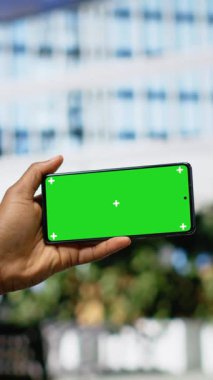 Dikey video yeşil ekran telefon ahşap bir bankta oturan adam, anlamlı tutum ve moda harmanlanmış çevre binalar, yol ayrıntıları ve aktif şehir hayatı