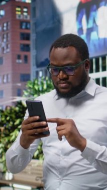 Dikey Video Siyah adam bir gezinti sırasında akıllı telefon harita uygulamasında yol tarifi arıyor, ofis binalarıyla şehir caddesinde yürüyor. Şehir merkezinde canlı bir şehir hayatı var.