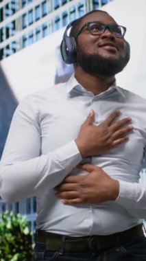 Dikey Video Trendi Afrikalı Amerikalı adam en sevdiği müzikle şarkı söylüyor ve dans ediyor, sokakta kulaklıkla harika dans hareketleri yapıyor. Kendine güvenen sokak tarzı ve şehir hayatının akışı. Kamera A.