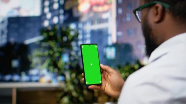 Şehir merkezindeki işlek bir caddede, şehir merkezinde gökdelenler ve dinamik manzarayla dolaşan siyahi bir adam tarafından tutulan dikey yeşil ekran telefon. Canlı bulvar. Kamera B.