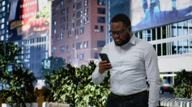 Afrika kökenli Amerikalı erkek şehir merkezindeki kaldırımda canlı kişiliğiyle gezinerek akıllı telefonunun GPS 'ini kontrol ediyor. Şehir hayatının modern dinamik mimarisi. Kamera B.