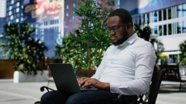 Şehir parkında bir bankta oturmuş dizüstü bilgisayar ve seyir sitesinde gezinen siyah bir adam, günışığı ve modern şehir manzarası ile vurgulanan günlük sokak tarzı ve davranışları. Kamera B.