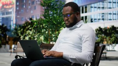 Afrikalı Amerikalı bir insan, ekrandaki kötü haberden hoşnutsuz ve hayal kırıklığına uğramış bir şekilde çevrimiçi çalışıyor. Metropolitan bölgesinde gökdelenlerle çevrili. Kamera B.