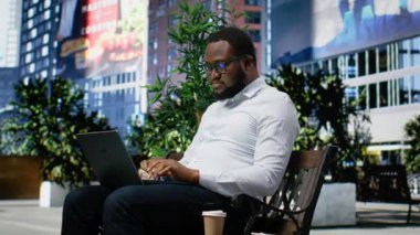 Siyah yetişkin bir adam dizüstü bilgisayarlı bir bankta oturmuş internet sitelerinde geziniyor, şehir merkezindeki küçük bir parkta güneş ışığının tadını çıkarıyor. Şehir içi hareketli modern şehir mimarisi. Kamera B.