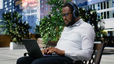 Uzaktan çalışma için odaklanmış müzik dinleyen kendine güvenen yetişkin bir adam, bir bankta oturuyor ve online işler için dizüstü bilgisayar kullanıyor. Şehir merkezinde modern bir caddede moda bir etkinlik. Kamera B.