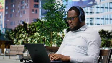 Afrikalı Amerikalı erkek, internet sitesinde dizüstü bilgisayarda geziniyor ve odak müziği dinliyor, parkta bir bankta oturuyor. Kalabalık bir şehir merkezinde metropoliten ufuk çizgisini ve şehirciliği çevrelemek. Kamera B.