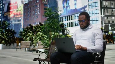 Afro-Amerikalı yetişkin bir erkek dizüstü bilgisayarında oturuyor, rahat yaşam tarzını ve iş odaklarını canlı mimariyle harmanlıyor. Şehir caddesinde büyük şehirler var. Kamera A.