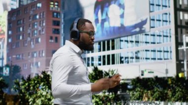 Afro-Amerikalı adam dans ediyor ve sokakta müzik dinliyor, kulaklık takıyor ve şehir kaldırımında neşeli hissediyor. Şık şarkıların ve şehir merkezinde imza günlerinin tadını çıkarıyorum. Kamera B.