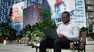 Afro-Amerikalı biri dışarıda park banklarında dizüstü bilgisayar kullanıyor, günlük sokak tarzıyla şehir bulvarı ve modern mimarinin ortasında uzaktan çalışmayı karıştırıyor. Kamera A.