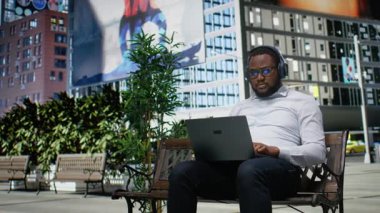 Afrikalı Amerikalı erkek, internet sitesinde dizüstü bilgisayarda geziniyor ve odak müziği dinliyor, parkta bir bankta oturuyor. Kalabalık bir şehir merkezinde metropoliten ufuk çizgisini ve şehirciliği çevrelemek. Kamera A.