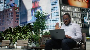 Parktaki bankta oturmuş video bağlantısı kuran siyahi bir adam online tartışma için dizüstü bilgisayarını kullanıyor ve şehir hayatının tadını çıkarıyor. Şehir merkezindeki modern sokak tarzı. Kamera A.