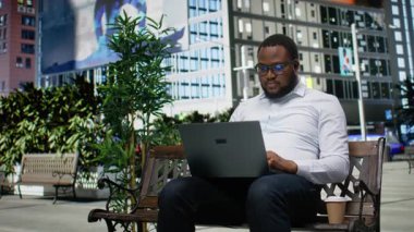 Parktaki bankta çalışan Afrikalı Amerikalı bir erkek, şehir merkezindeki bir bankta e-posta yazıyor ve içeriği okuyor. Bulvar yakınlarında gökdelenler ve metropoliten çevre. Kamera A.