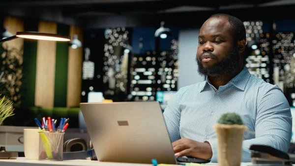 Çok uluslu bir şirket için e-posta okuyan ve web bilgilerini belgeleyen siyah bir çalışan karanlık bir ofiste saatler sonra çalışıyor. Girişim hedefleri ve gelişimi.