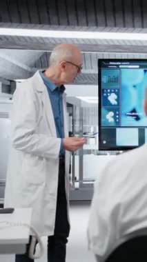 Dikey video üst düzey bilim adamı laboratuardaki ilaç geliştirme ekibine yeni bulgular sunuyor. Antrenman sırasında hücresel reaksiyon gösteren büyük ekrana bakan uzman, kamera B