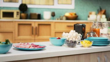 Modern bir tezgahta pizza malzemeleriyle donatılmış rahat bir mutfak. Peynir, domates ve biber kâseleri baharat ve mutfak malzemelerinin yanında oturur, eğlenceli bir hafta sonu yemeği aktivitesine hazırdır..