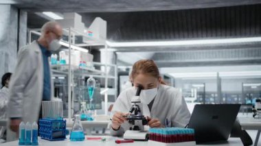Tıbbi laboratuvarda cerrahi maske takan bir biyolog hücresel analiz için bilimsel mikroskop kullanıyor. Laboratuvarda yüksek teknoloji optik cihaz teçhizatı ve kamera A ile birlikte kadın bilim adamı örneği inceliyor.