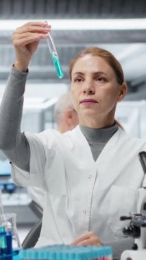 Dikey video Kalite Kontrol Analisti test tüpündeki mavi sıvı berraklığı inceleyip laptopa veri girişi yapıyor. Laboratuvar teknisyeni endüstriyel ürün üretimi için kimyasal saflık standartlarını doğruladı