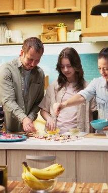 Dikey Video Ailesi, lezzetli ev yapımı pizzalar yaptıktan sonra el çırpıyor, sıcak bir mutfakta tezahürat yapıyor ve alkışlıyor. Ebeveynler ve küçük kız çocukları kahkaha ve sıcaklığı paylaşıyorlar. Kamera A.