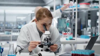 Mikroskop inceleyen dişi mikroskopist hücreleri görmek için laboratuarda cam kaydırma yapıyor. Araştırma tesisi çalışanı örnek slaytı kontrol eder, optik aygıt kullanarak görüntüleme işlemi yapar, kamera B