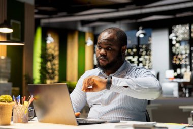 Siyah ofis çalışanı kol saatini kontrol ediyor ve geç saatlere kadar çalışıyor. Acele edip gece yarısı görevlerini bitirmeye çalışıyor. Yorgun işadamı ofiste fazla mesai yapıyor..