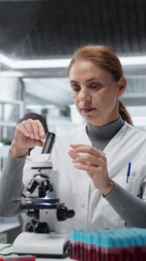 Laboratuvarda detaylı hücresel analiz araştırmaları için bileşik mikroskop kullanan dikey video moleküler biyolog. Kadın kalite kontrol analisti modern laboratuvarda doku örneği incelemesi yapıyor.