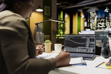 Siyah finans danışmanı, kaynak tahsisini planlamak ve ticari hedefleri mali başarı ile sıralamak için dijital grafikler kullanarak, gelecekteki raporlar için ticaret hesapları ve arşivlenmiş verileri gözden geçirir.