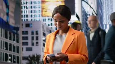 Siyah modern profesyonel kadın akıllı telefonunu kontrol ediyor ve şehir merkezinde yürüyor, sokak insanları ve şehir kalabalığıyla çevrili, günlük hareketlerini ve iş rutinlerini ifade ediyor. Kamera B.