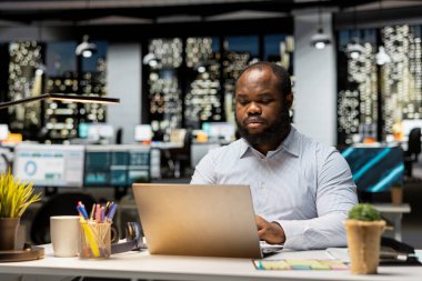 Çok uluslu bir şirket için e-posta okuyan ve web bilgilerini belgeleyen siyah bir çalışan karanlık bir ofiste saatler sonra çalışıyor. Girişim hedeflerine ve gelişimine odaklanır.