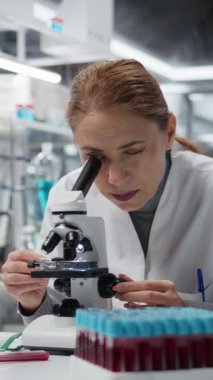 Dikey video Histoloji teknisyeni mikroskop altında patoloji slaytları inceliyor, çalışma tezgahındaki kötü duruşu düzeltiyor. Tıbbi yenilikleri hücresel analiz yoluyla yürüten araştırmacı sırt ağrısı hissediyor.