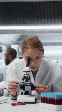 Mikroskop incelemesi yapan dikey video kadın mikroskopist laboratuarda hücreleri görmek için cam kaydırağını inceliyor. Araştırma tesisi çalışanı örnek slaytı kontrol eder, optik cihaz kullanarak görüntüleme işlemi yapar, kamera A