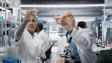 Yeni ilaç formülasyonu için bileşik çözümleri değerlendirmek üzere laboratuvarda staj öğreten kıdemli bir araştırmacı. Kadın bilim adamı ve yaşlı laboratuvar çalışanı deney ilaçlarını şişede test ediyorlar, kamera B