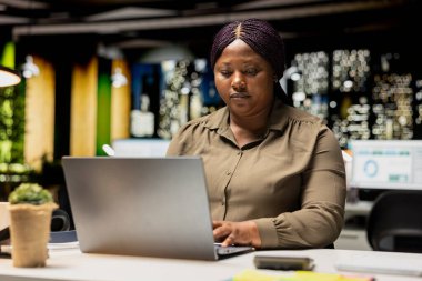 Afrikalı Amerikalı analist e-postaları okuyor ve yönetimsel görevleri yönetiyor. Çok uluslu bir şirket için güncellemeleri belgeliyor. Gece vakti karanlık bir ofiste iş hedefleri üzerinde çalışıyorum..