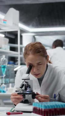 Bilimsel mikroskobu hücresel analiz için kullandıktan sonra, dikey video bitkinliği çeken biyolog tıp laboratuarında geriliyor. Yorgun bilim adamı çalışma masasında dinleniyor, optik cihaz teçhizatı ile numuneleri inceliyor.