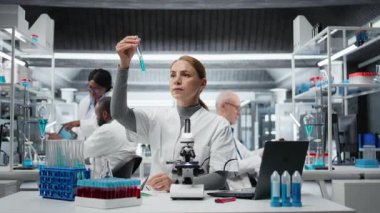 Kalite kontrol analisti test tüpündeki mavi sıvı berraklığı inceliyor ve laptopa veri giriyor. Endüstriyel ürün üretim süreci için kimyasal saflık standartlarını onaylayan laboratuvar teknisyeni, kamera A