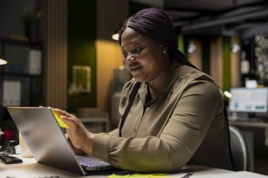 Siyahi girişimci kadın, ertesi sabah önemli bir görevi hatırlamak için bir not kullanarak dizüstü bilgisayarına yapışkan notlar yapıştırıyor. İşlerini gece geç saatlerde ofiste bitiriyor..