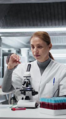 Laboratuvardaki dikey video tarih teknisyeni, yapısal bozukluklar ya da safsızlıklar için ince cam kaydırağını inceliyor. Laboratuvarda çalışan bir kadın mikroskop kullanarak görsel kalite kontrolü yapıyor.