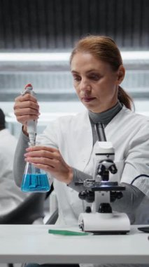 Dikey video moleküler biyolog saflık analizi için mavi çözeltiye tepki veriyor. Orta yaşlı bir kadın kimyasal reaksiyon özelliklerini gözlemlemek için test tüpüne sıvı madde ekliyor. Kamera A.