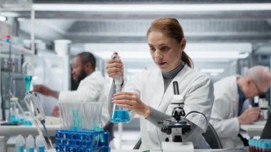 Laboratuvar geliştirme uzmanı test tüpüne mavi sıvıyı koyup analiz ediyor. Moleküler biyolog, spektroskopik analize hazırlanmak için kimyasal örnek aktarıyor. Kamera B.