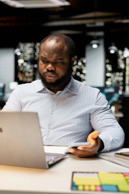 Siyahi işadamı cep telefonundan iç çekip mesajları kontrol ediyor, modern ofisteki bir tebligat yüzünden dikkati dağıldıktan sonra tükenmiş ve kaşlarını çatmış hissediyor. Geceleri işyeri sorunları.