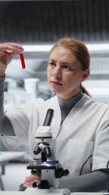 Hastalık araştırmaları klinik testleri için kan örneklerini analiz eden dikey video laboratuarı uzmanı. Laboratuvarda test için sanguin sıvısı şişeleri üzerinde araştırma yapan kadın bilim adamı, kamera A