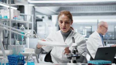 Mikrobiyolog bakteri kolonisinin büyümesini kontrol etmek için petri kabını ışığa doğru tutuyor. Görünür kirlenme belirtileri için tarım levhası kültürünü inceleyen kadın teknisyen, kamera B