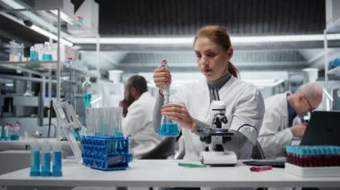 Moleküler biyolog saflık analizi için mavi çözeltiye tepki veriyor. Orta yaşlı bir kadın kimyasal reaksiyon özelliklerini gözlemlemek için test tüpüne sıvı madde ekliyor. Kamera A.