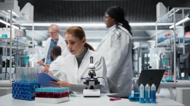 Laboratuvar çalışanı mikroskop ile biyopsi slaytını inceliyor, dosyada teşhis bulgularını belgeliyor. Orta yaşlı bir kadın klinik laboratuarında hastalık belirtilerini tespit ediyor, not alıyor, kamera A