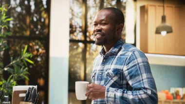 Afro-Amerikalı neşeli bir adamın portresi kahvaltıda ekmek ve kruvasan yemeden önce sabah kahvesini fincanda içiyor. Mutfak eşyaları ve sağlıklı yiyeceklerle çevrili sakin bir sabah..