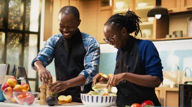 Siyah erkek arkadaş salataya baharat ve baharat ekliyor, organik öğle yemeğini taze biyolojik malzemelerle tatlandırıyor. Kız arkadaşın sebzeleri bir kasede karıştırıyor, vejetaryen yemeği..