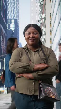 Kavşakta kırmızı ışıkta bekleyen, kalabalık bir şehir kalabalığıyla kalabalık metropolitan bölgesinde yürüyen Afro-Amerikan sıradan bir kadının dikey video portresi. Farklı alışkanlıkları olan çok ırklı insanlar.