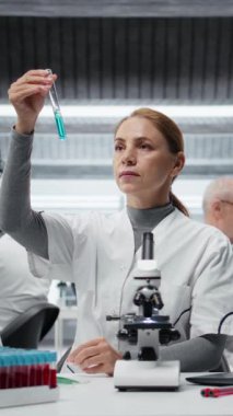 Dikey video Kalite Kontrol Analisti test tüpündeki mavi sıvı berraklığı inceleyip laptopa veri girişi yapıyor. Laboratuvar teknisyeni endüstriyel ürün üretimi için kimyasal saflık standartlarını doğruladı