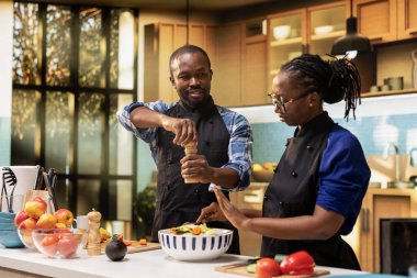 Siyah erkek arkadaş salata çeşnili biber ve kız arkadaş onu durduruyor. Sağlıklı bir öğle yemeği için yeterli baharat ve baharat var. Önlüklü mutlu çift birlikte yemek hazırlıyor..